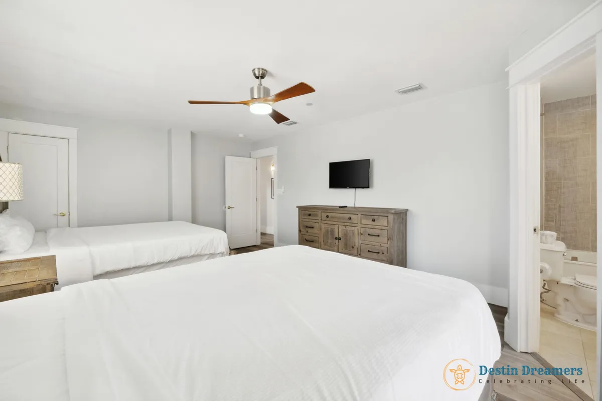A welcoming double queen suite, featuring two large beds with soft linens, a wooden dresser, ceiling fan, and a wall-mounted smart TV. The room has a soothing color scheme and is designed for comfort and shared relaxation, ideal for teens or friends traveling together.