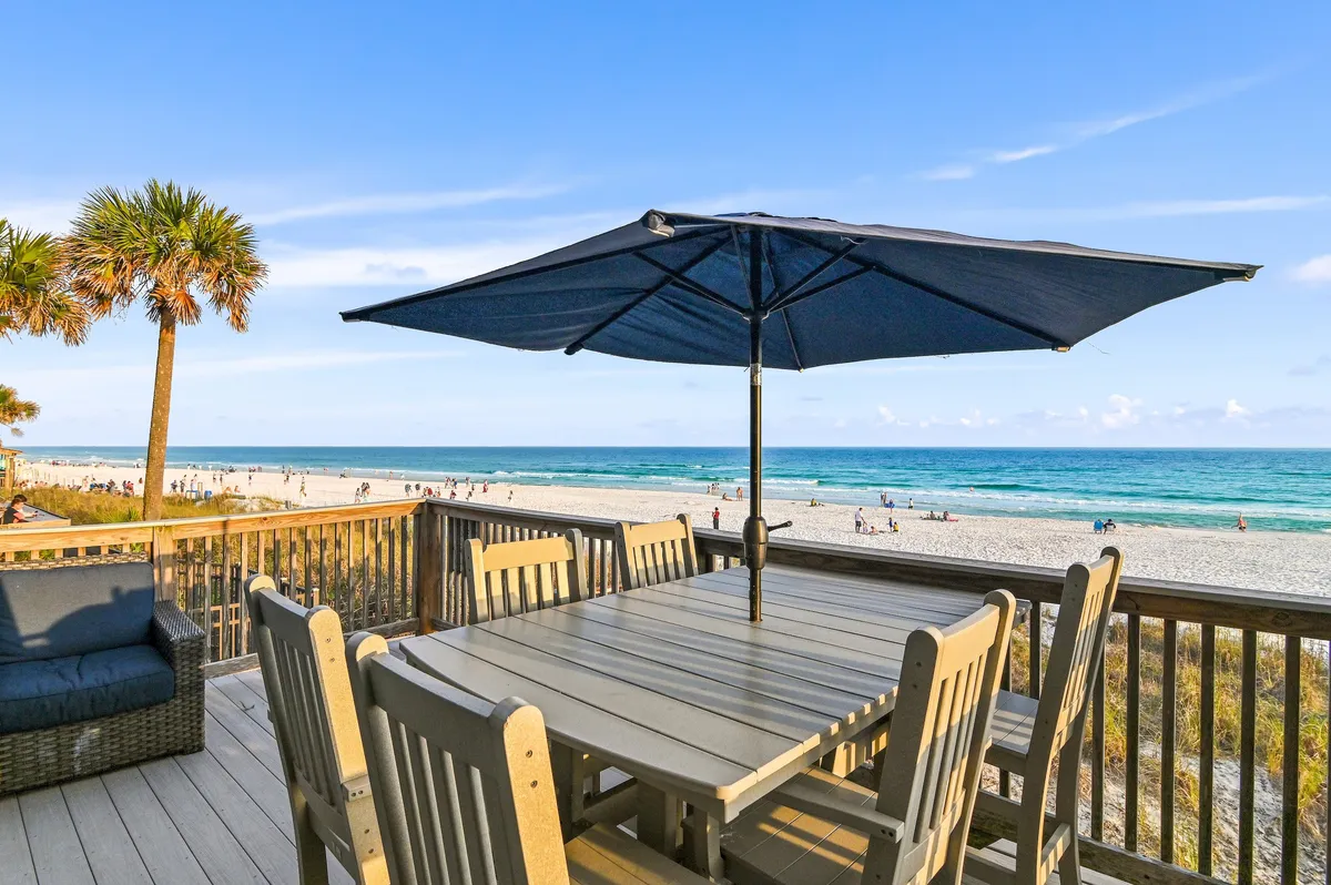 The hero image showcases a spacious wooden deck with a stylish outdoor dining table shaded by an umbrella, cozy lounge seating, and sweeping views of the pristine white sand beach and sparkling Gulf waters. A palm tree adds a tropical vibe, and the private deck leads directly to the uncrowded shoreline, suggesting exclusive beach access and the perfect spot for dolphin watching at sunset.
