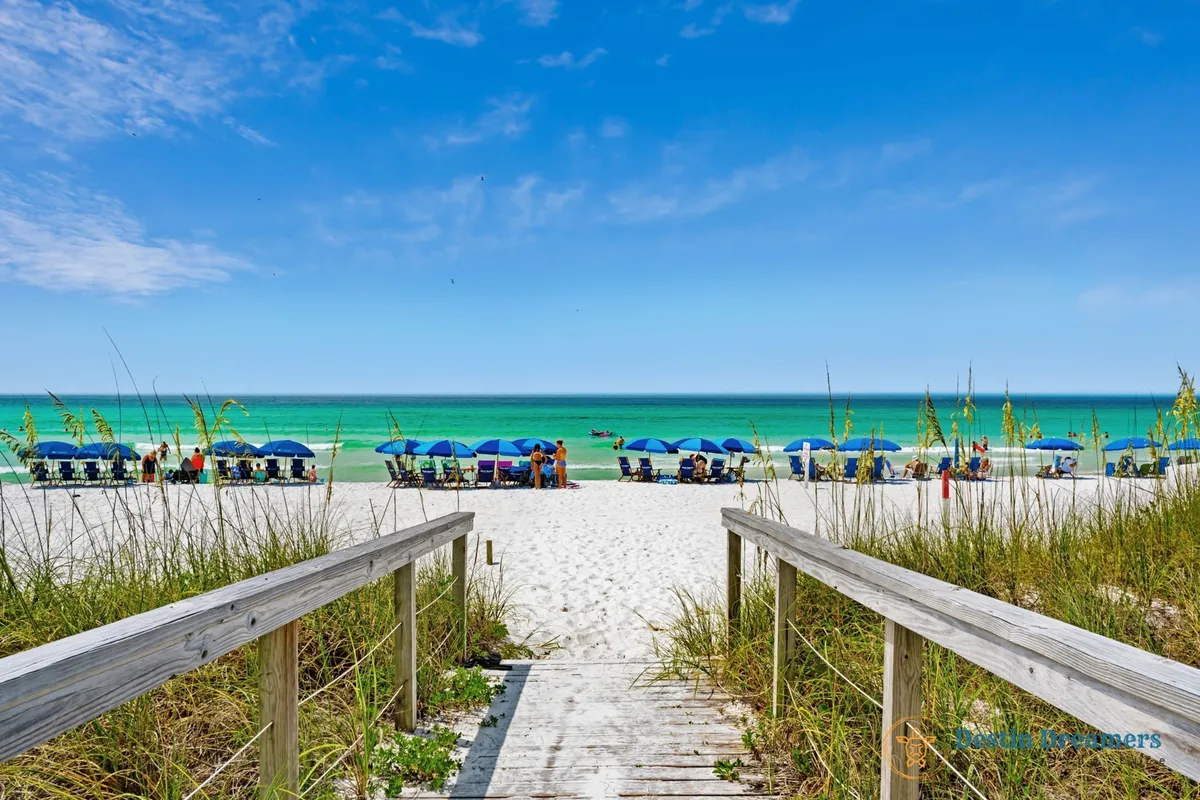 A wooden boardwalk leads from the property to a stretch of white sandy beach lined with blue umbrellas and chairs. The turquoise Gulf sparkles in the background, and the area is notably uncrowded, emphasizing privacy and tranquility.