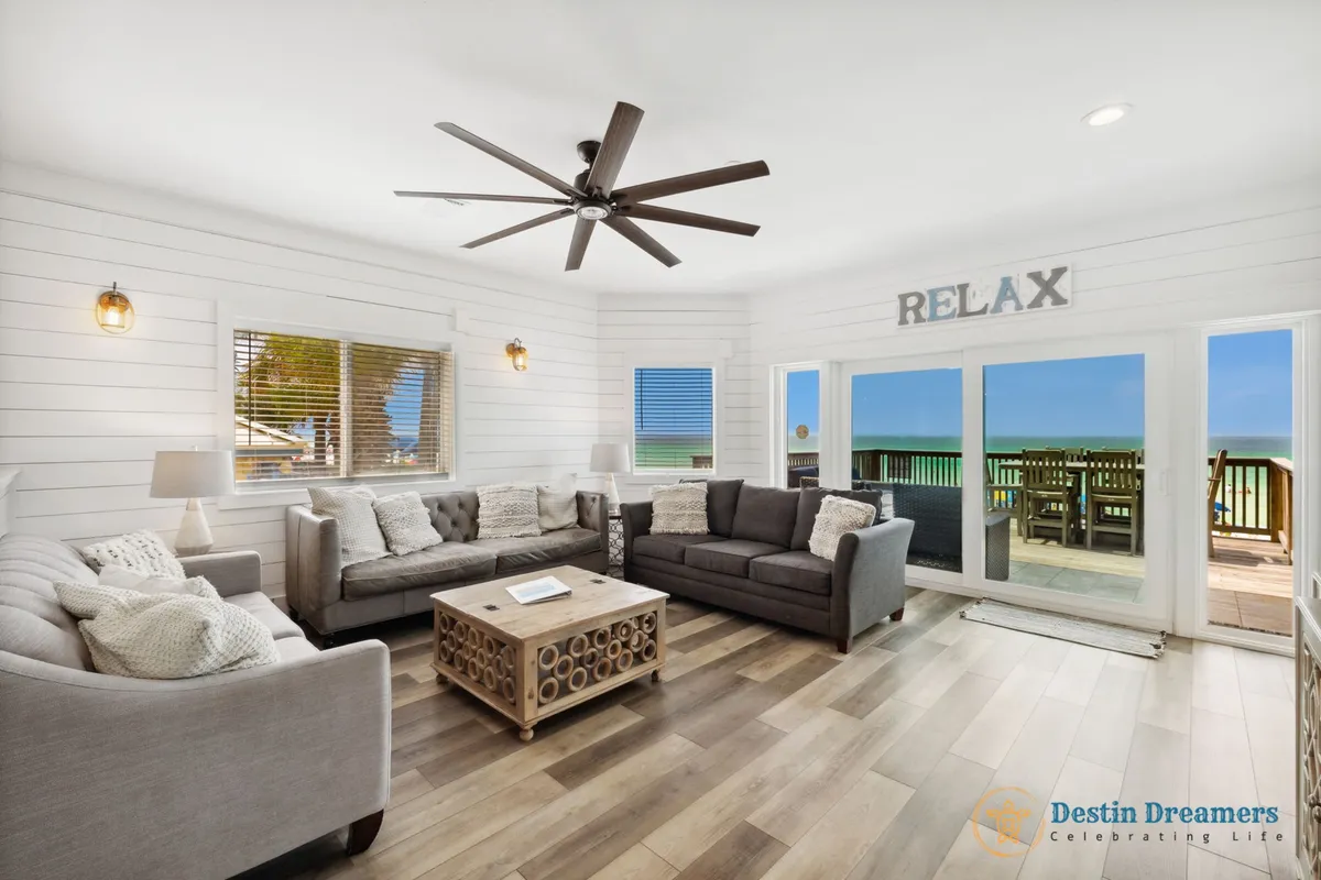 A spacious living room with three sofas arranged for conversation, a wooden coffee table, and large sliding glass doors that open to a balcony with panoramic Gulf views. The room is bright and airy, with a 'RELAX' sign above the doors, creating an inviting atmosphere perfect for unwinding after a day at the beach.