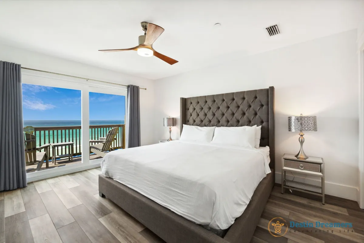 A spacious king master suite featuring a large tufted headboard, floor-to-ceiling sliding glass doors that open to a private balcony with seating overlooking the Gulf, and a modern ceiling fan. The room is filled with natural light and offers seamless indoor-outdoor living for relaxation and sunset views.