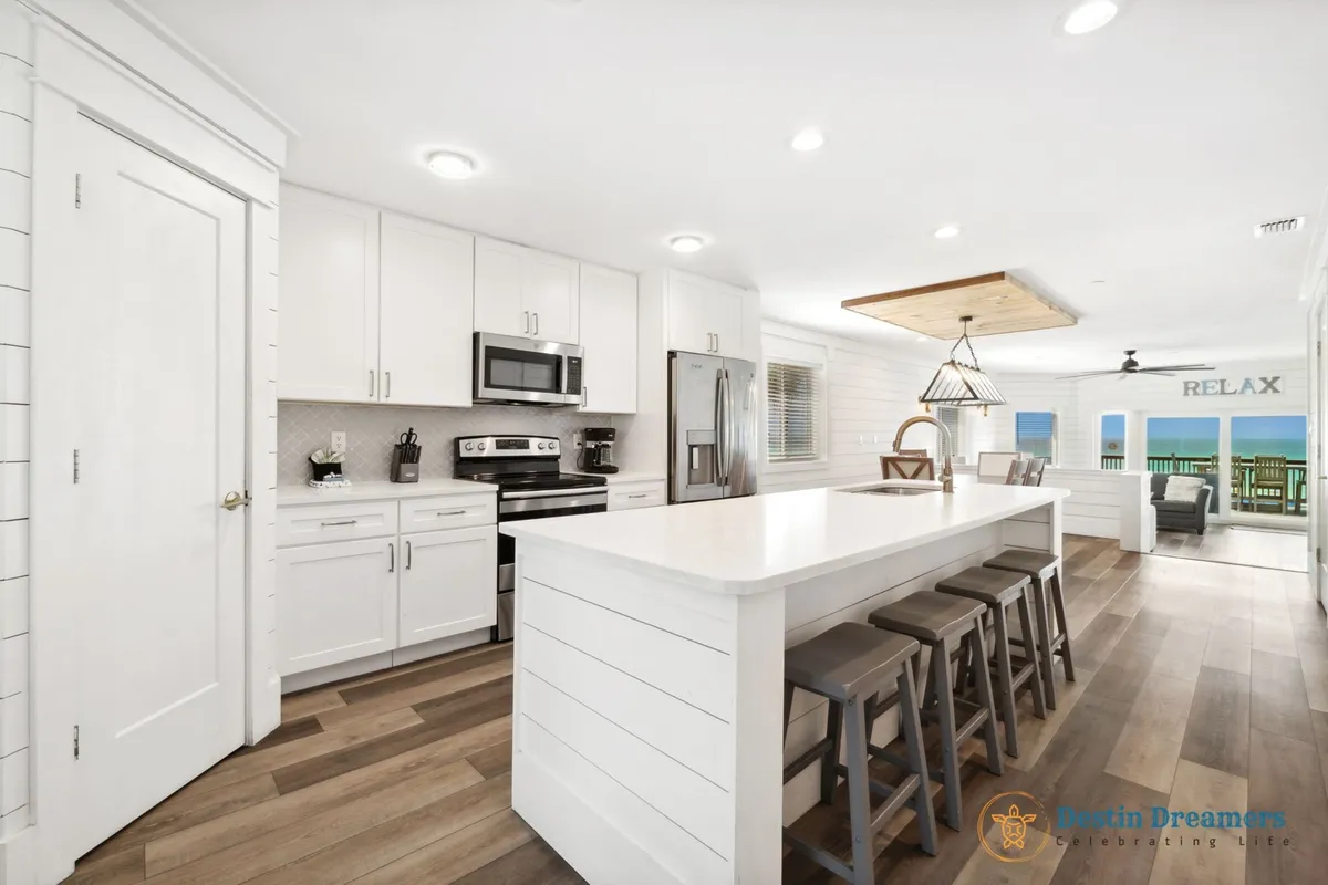 A spacious, open-plan kitchen features a large white island with bar seating, stainless steel appliances, and a decorative light fixture. The layout flows seamlessly into the living area and towards sliding glass doors that open to a balcony with a gorgeous ocean view, all bathed in natural light from floor-to-ceiling windows.