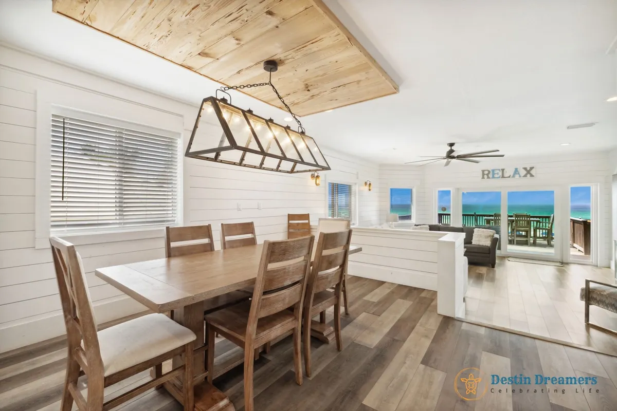 A wooden dining table with six chairs is set in front of a large window with a direct ocean view. The adjacent living area includes cozy seating and sliding glass doors that lead out to the balcony. A decorative 'RELAX' sign adds a welcoming touch.