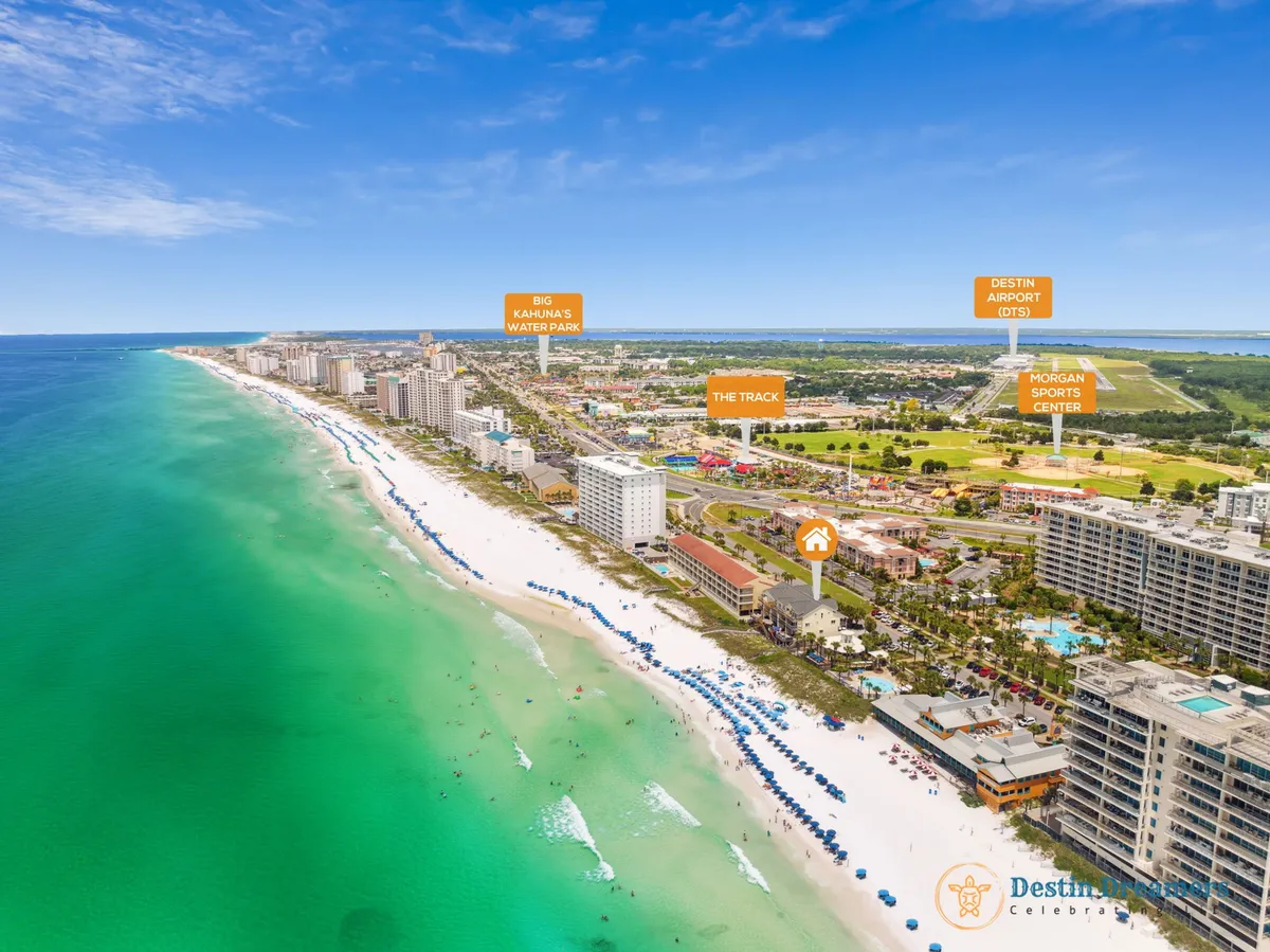 A wide aerial view of Destin’s white sandy beach lined with rows of blue umbrellas and chairs, turquoise Gulf waters stretching out, and beachfront buildings and resorts, with the featured property highlighted. The lively shoreline hints at water sports and bustling activity, while the ocean beckons just steps away.