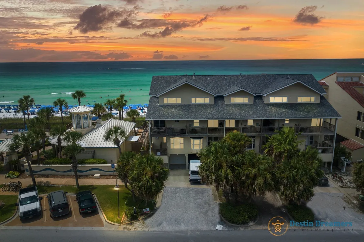 A multi-story beachfront townhouse with private balconies overlooks a pristine stretch of white sand and turquoise Gulf waters. The scene is bathed in vibrant orange and pink sunset hues, with blue beach umbrellas, palm trees, and lush tropical landscaping enhancing the exclusive, serene vibe. Parked vehicles in the foreground hint at easy access for guests.