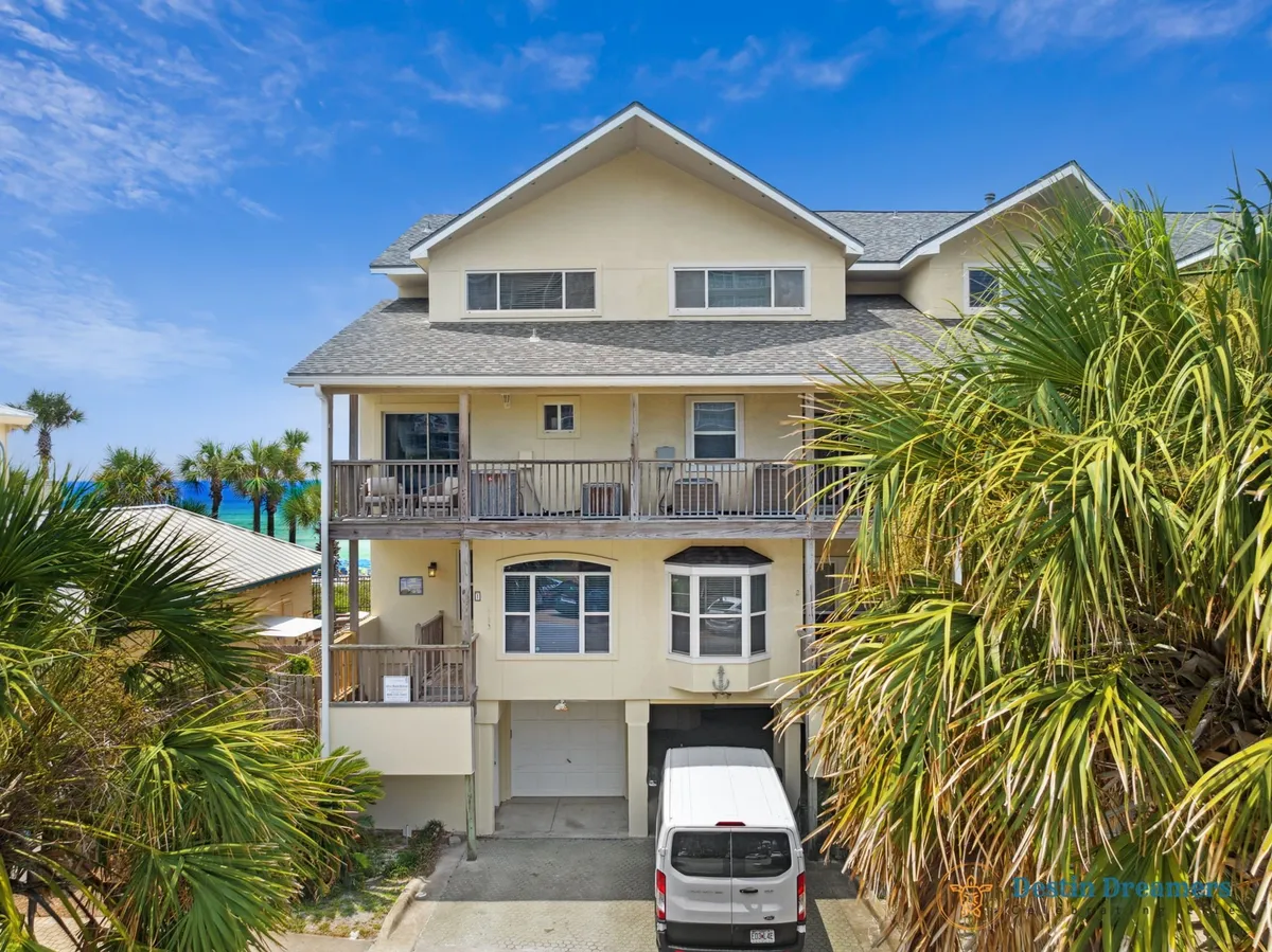 The three-story townhouse with multiple balconies overlooks the Gulf, framed by palm trees and a clear blue sky. The driveway and parked van convey readiness for excursions, while the balconies suggest relaxation and ocean views after a day of adventure.