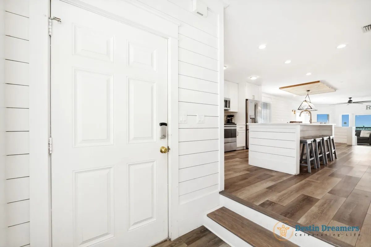 A spacious and bright interior scene featuring a modern kitchen island with wooden stools, stainless steel appliances, and a welcoming front door. Large windows in the living area bring in natural light and hint at outdoor access, creating a seamless transition between inside and out.