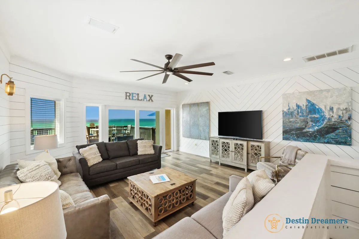 A cozy living room with three sofas and textured pillows is framed by large windows and a sliding door, offering vibrant turquoise ocean views. A unique wooden coffee table and the 'RELAX' sign above the door create a welcoming, beachy vibe.