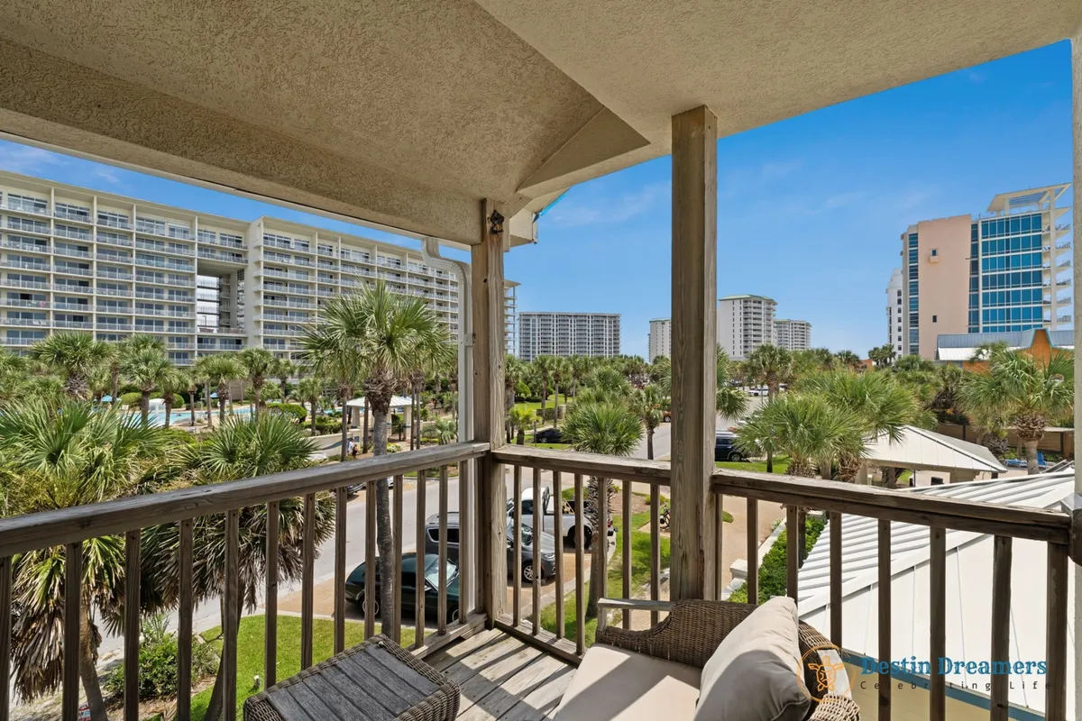 An inviting exterior shot of a wooden balcony with comfortable seating, overlooking palm trees and nearby resorts under a clear blue sky. The perspective suggests a peaceful spot to relax and observe the neighborhood, blending privacy with a sense of place.