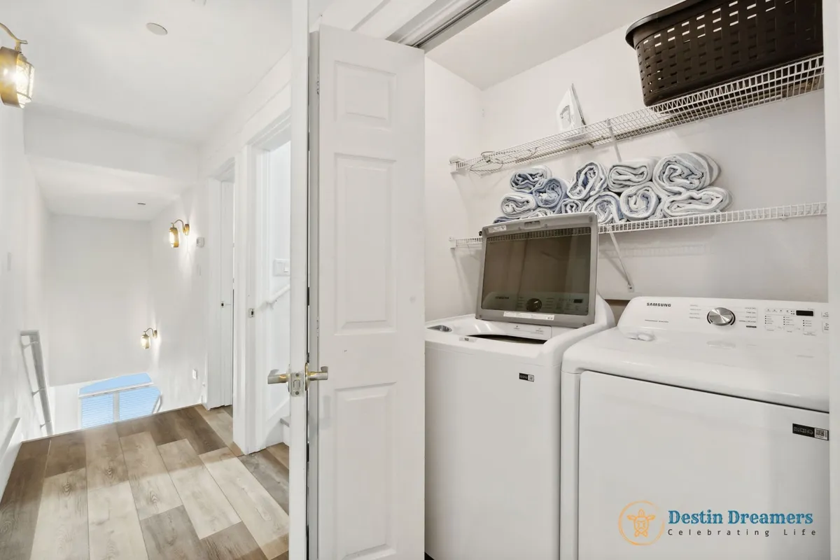 A dedicated laundry area with washer and dryer machines, open shelving with rolled towels, and a bright hallway connecting bedrooms. The setup is tidy and practical, enhancing convenience for extended stays.
