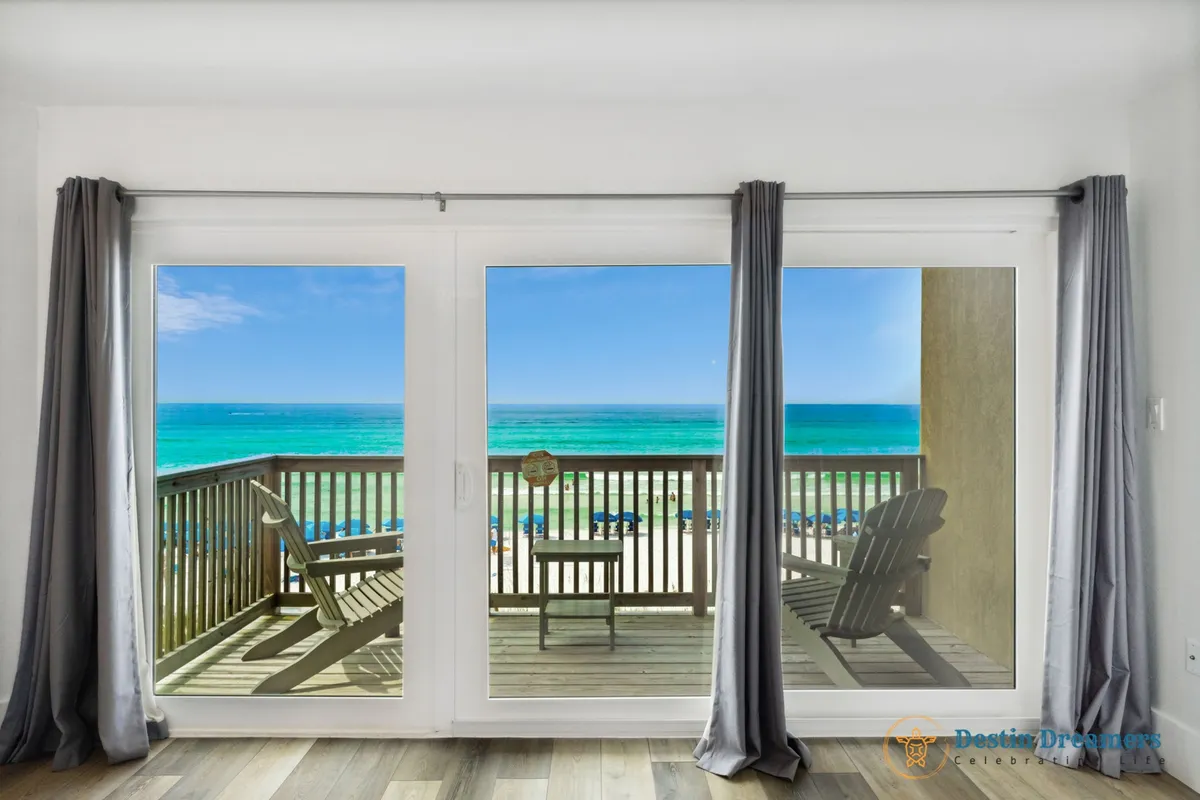 Large sliding glass doors open to a private balcony furnished with Adirondack chairs and a small table, framing breathtaking turquoise Gulf waters and a clear blue sky. The view is expansive, offering a sense of tranquility and connection to the coast.