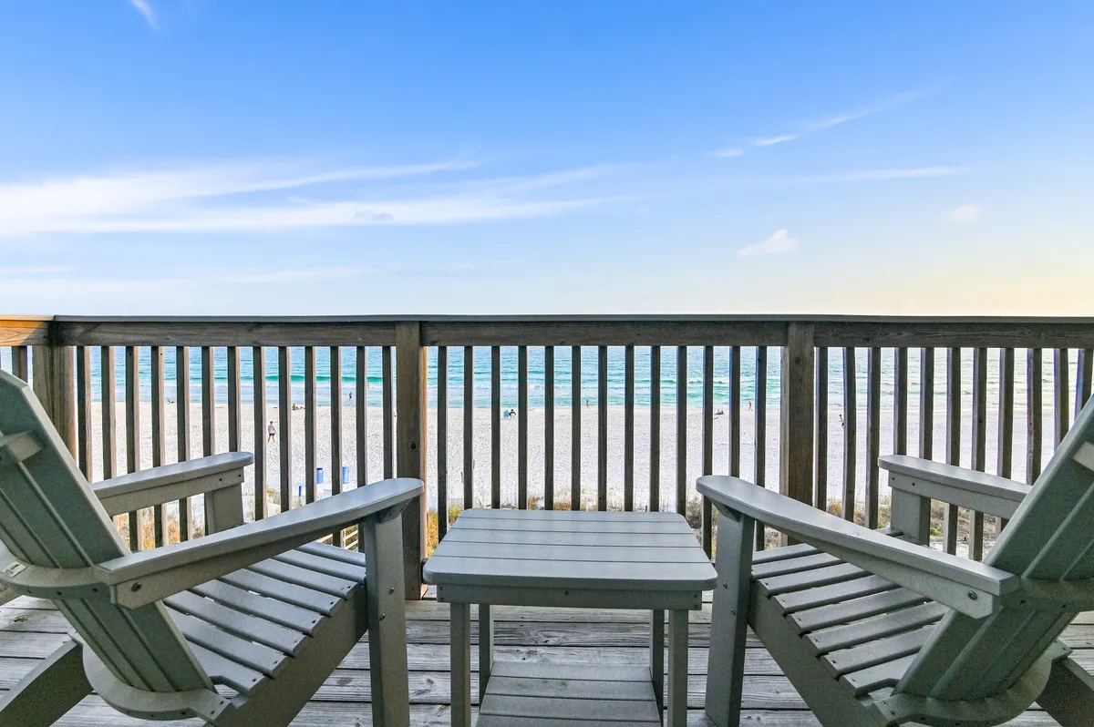 Two Adirondack chairs on a wooden balcony overlook the white sandy beach and calm turquoise Gulf waters. The setting is peaceful and intimate, perfect for relaxing with loved ones or enjoying a sunset with a pet at your feet.