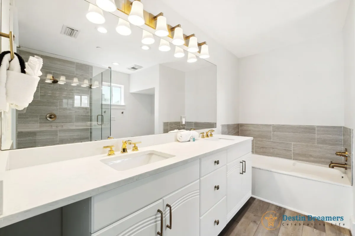 A luxurious, spacious bathroom featuring a double vanity with gold fixtures, a large mirror with bright lighting, a glass-enclosed shower, and a bathtub with tiled surround. The space is bright, modern, and designed for relaxation.