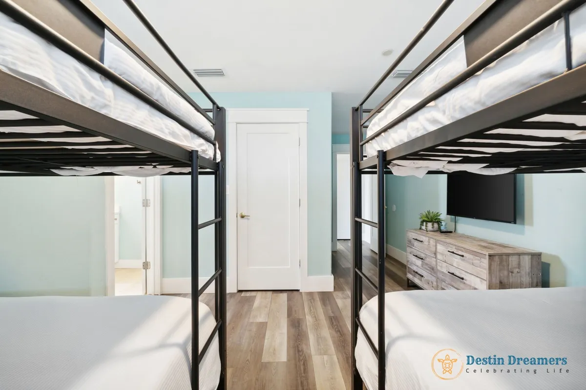 A cheerful bunk room with two sets of sturdy black metal bunk beds, light blue walls, a wooden dresser with a potted plant, and a wall-mounted flat screen TV. The space is bright, organized, and clearly designed for groups or families with children.