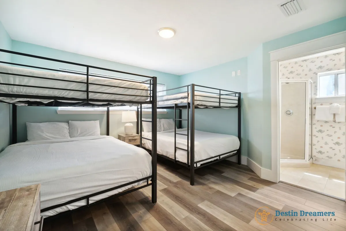 A bright, comfortable guest suite with two black metal bunk beds (queen on bottom, twin on top), light blue walls, a wooden nightstand with lamp, and an open door leading to a private bathroom. The space is family-friendly and designed for group comfort.