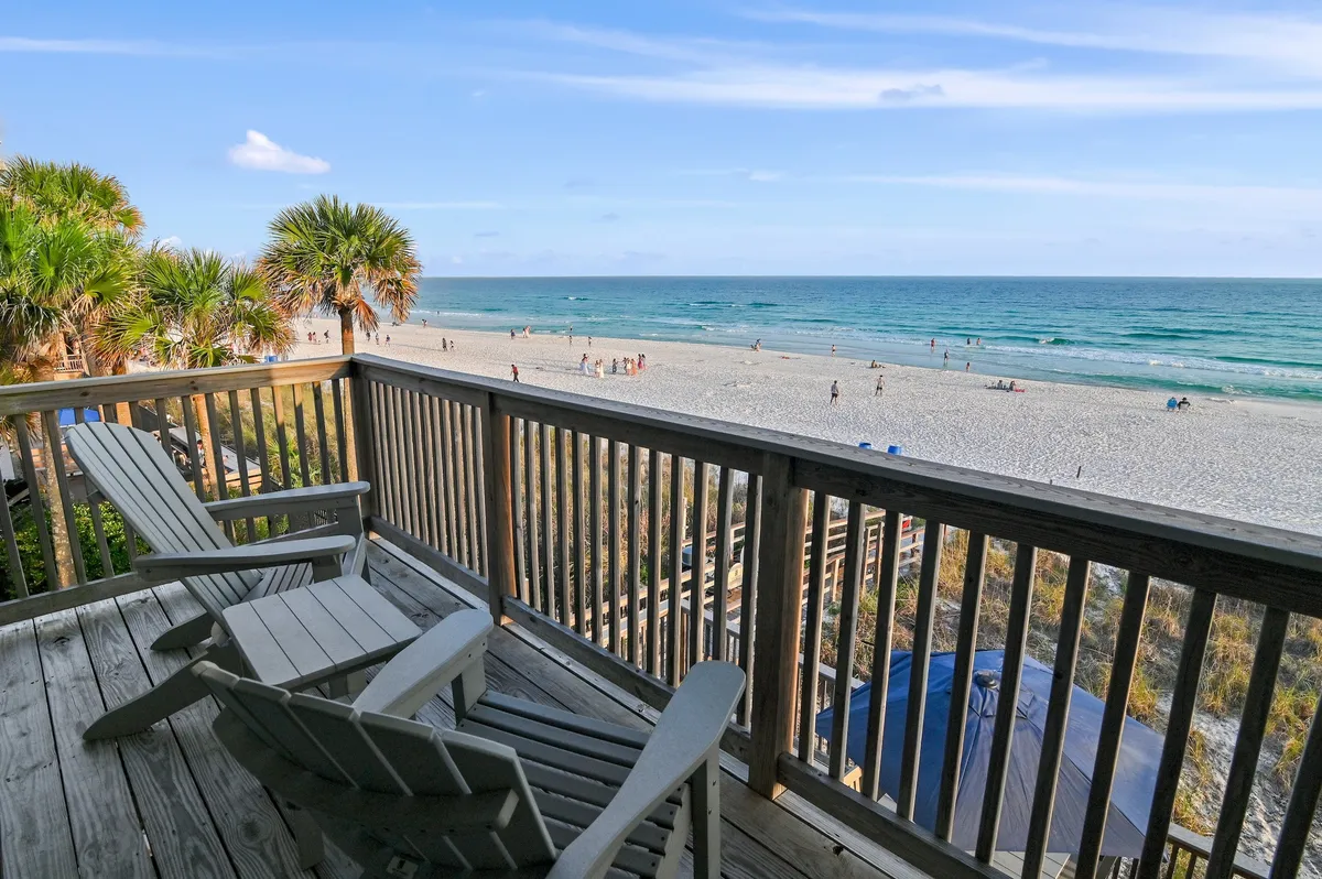 A wooden balcony furnished with Adirondack chairs overlooks a pristine, uncrowded white sand beach and the shimmering turquoise Gulf. The scene radiates calm and privacy, with only a few distant beachgoers, suggesting an exclusive and peaceful coastal retreat.