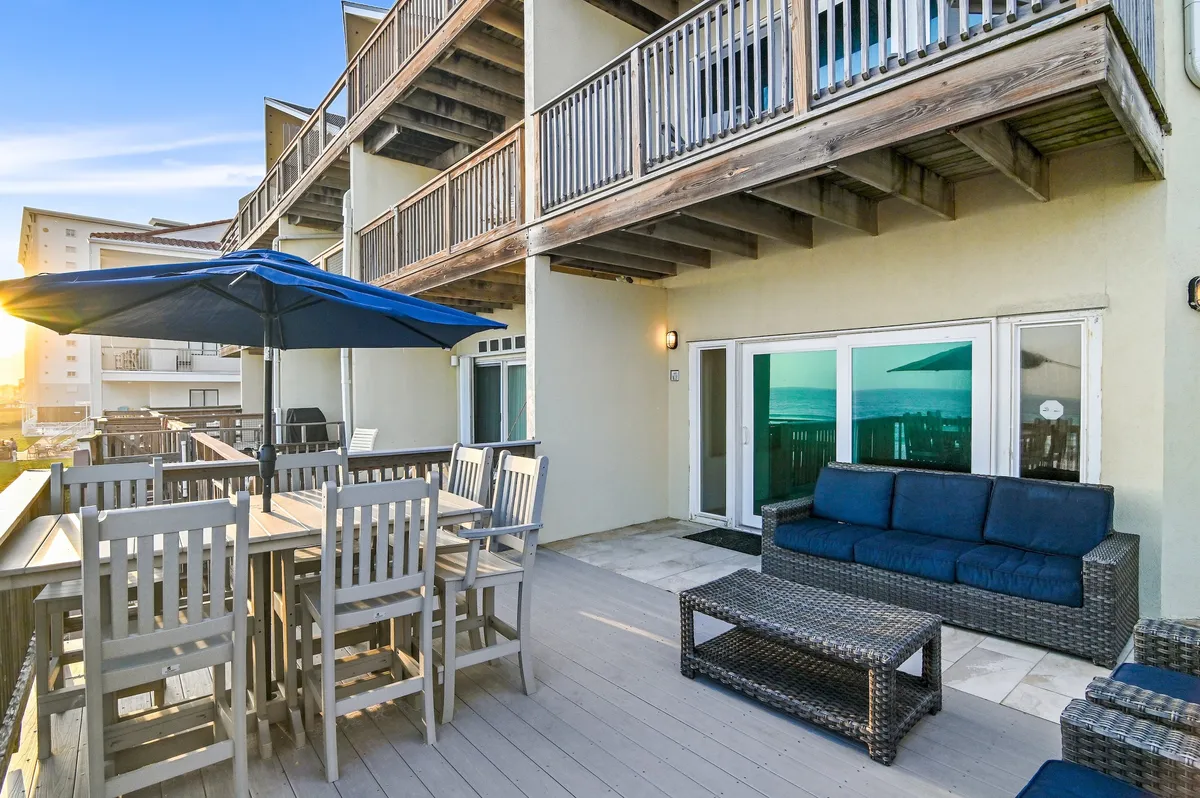 A wooden balcony features an outdoor dining table with an umbrella, wicker sofa seating with blue cushions, and sweeping views of the ocean. Sliding glass doors reflect the Gulf, emphasizing the blend of indoor comfort and outdoor enjoyment.