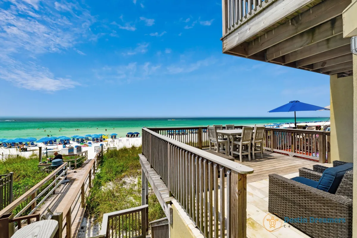 A wooden deck set for outdoor dining with a table and chairs shaded by a blue umbrella, overlooking turquoise Gulf waters and a white sandy beach. In the distance, people are relaxing under rows of matching blue umbrellas, and a wooden pathway leads directly to the sand.