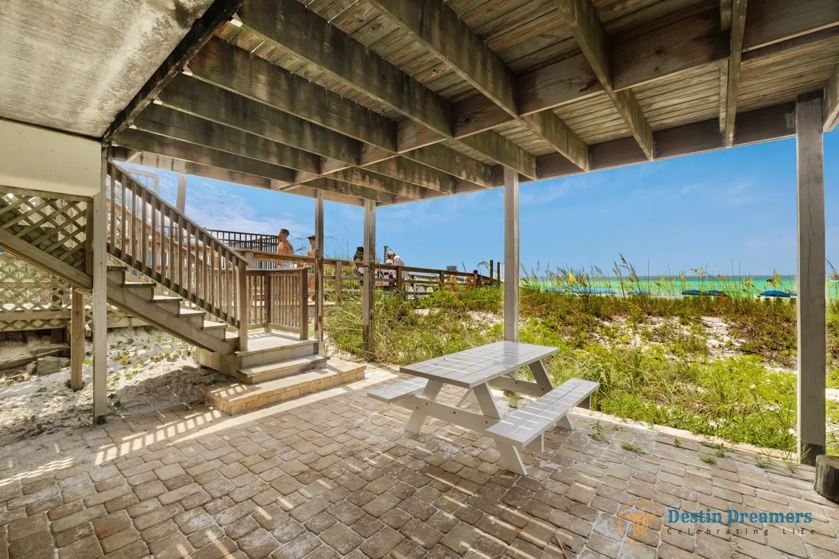 This exterior image features a wooden staircase leading directly to a sandy beach, with a shaded picnic table under the deck and tranquil turquoise Gulf waters in the background. Dune grass frames the path, and the scene captures the essence of private, easy beach access in a serene setting.