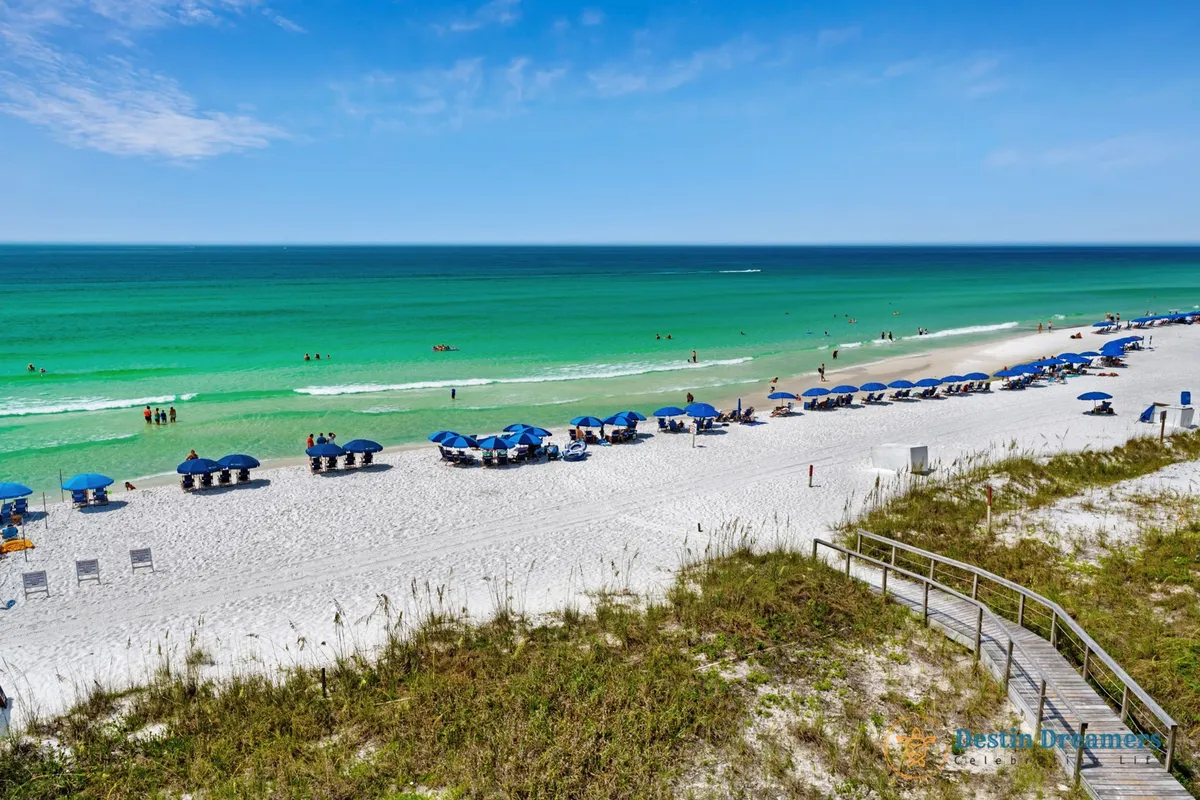 A tranquil view of the private white sandy beach with rows of blue umbrellas and chairs, turquoise Gulf waters, and a wooden walkway over the dunes. The scene suggests both relaxation and the starting point for beach activities, water sports, or dolphin watching.