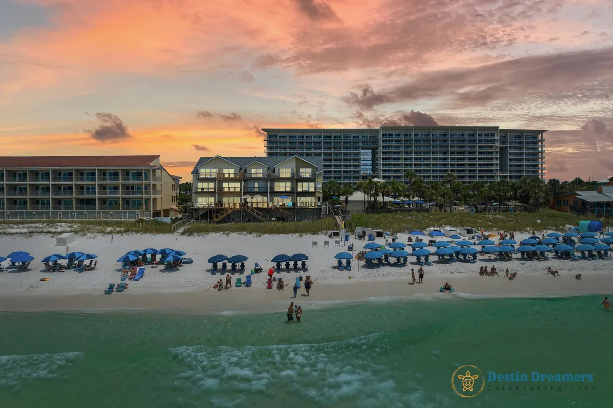A row of exclusive beachfront townhouses, each with private balconies overlooking a pristine, uncrowded beach. Blue umbrellas and chairs are organized along the sand, with people enjoying sunbathing, swimming, and strolling by the water. The sky is painted with warm sunset colors, enhancing the feeling of tranquility and luxury.