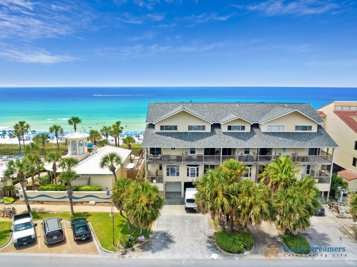 A multi-level beachfront townhouse with private balconies overlooking the Gulf of Mexico, pristine white sand, and rows of umbrellas and palm trees. The property feels exclusive and modern, with easy access to the beach and amenities like parking and bikes for guests.