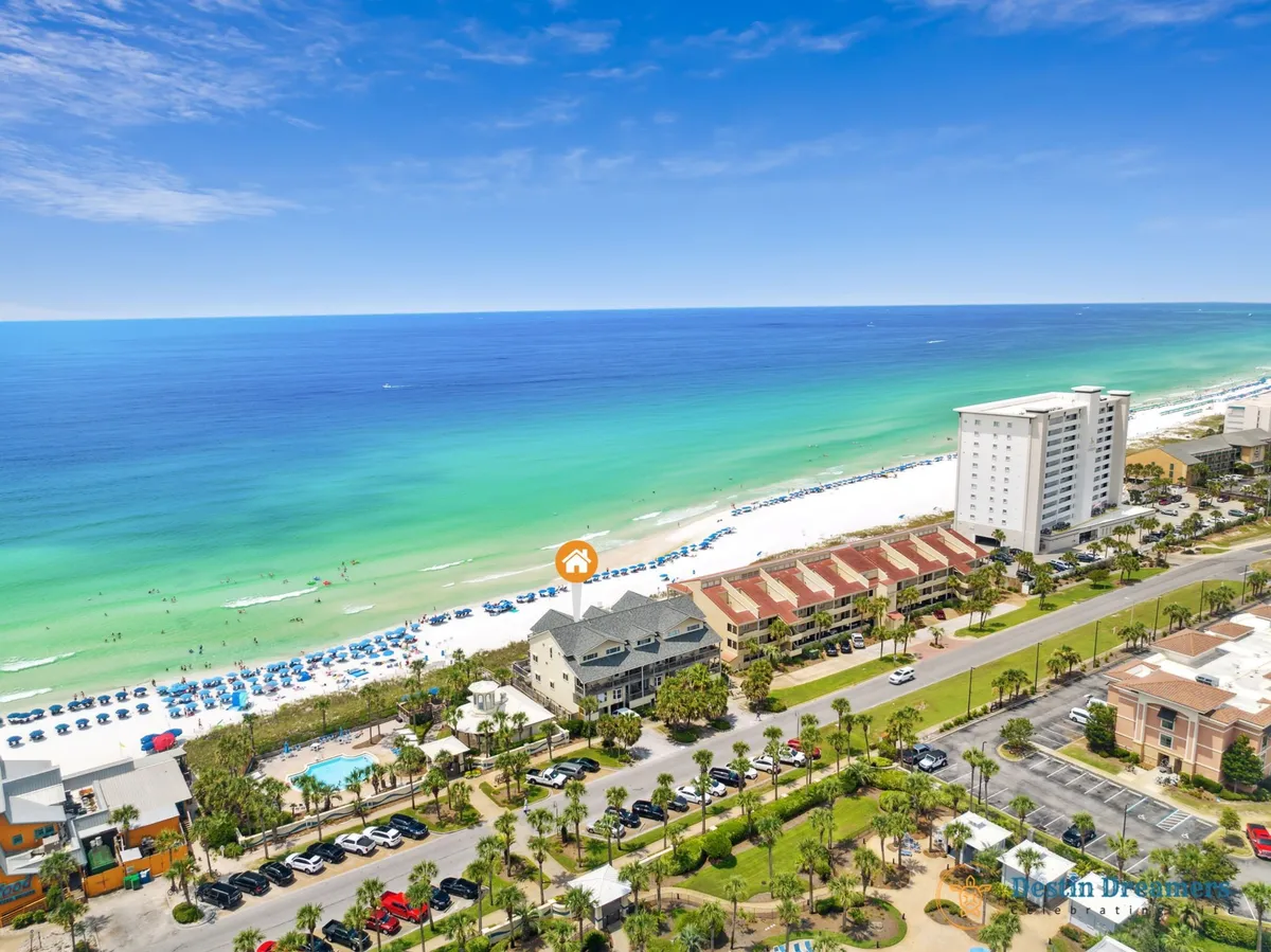 A sweeping aerial view reveals the vast turquoise Gulf meeting white sandy shores, lined with neat rows of beach umbrellas and distinctive townhouse buildings nestled among palm-lined streets. The sense of openness, privacy, and connection to both the beach and the exclusive property is palpable.